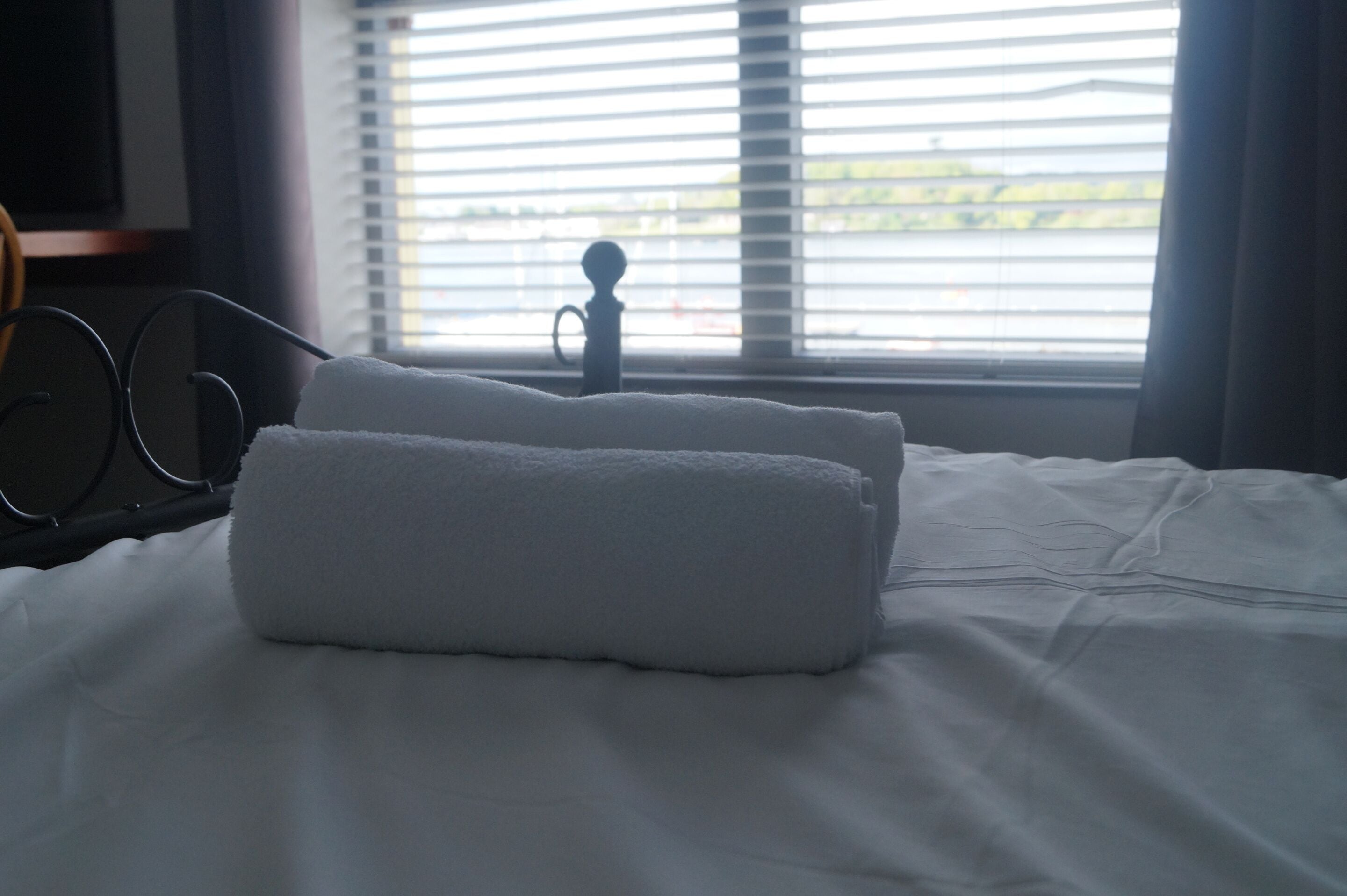 A neatly made bed with a folded white towel, a pair of glasses on a stand, and a window with blinds.