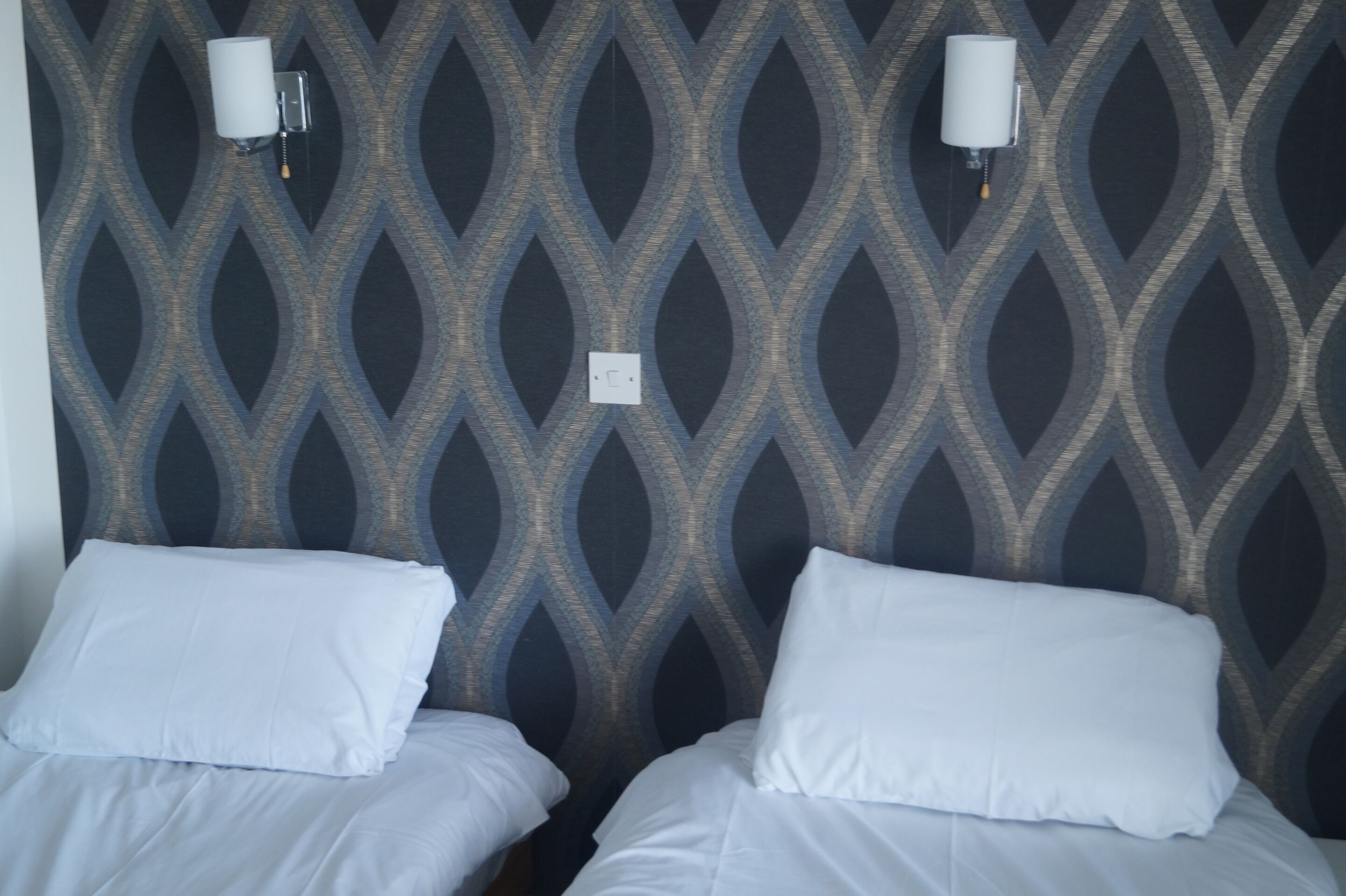 Habitación con dos camas, un papel tapiz estampado y dos luces fijadas a la pared.