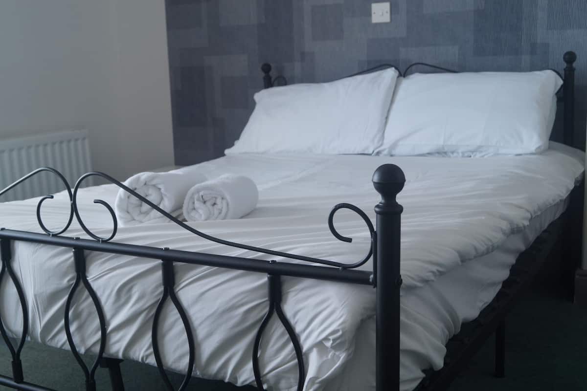 Un lit bien fait, avec du linge de lit blanc et une serviette enroulée posée sur le côté, une tête de lit en métal noir et un mur à motifs en arrière-plan.