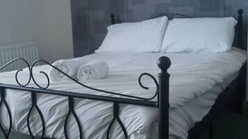 Una cama bien hecha con sábanas blancas y una toalla enrollada al lado, cabecera de metal negro y un papel tapiz estampado en el fondo.