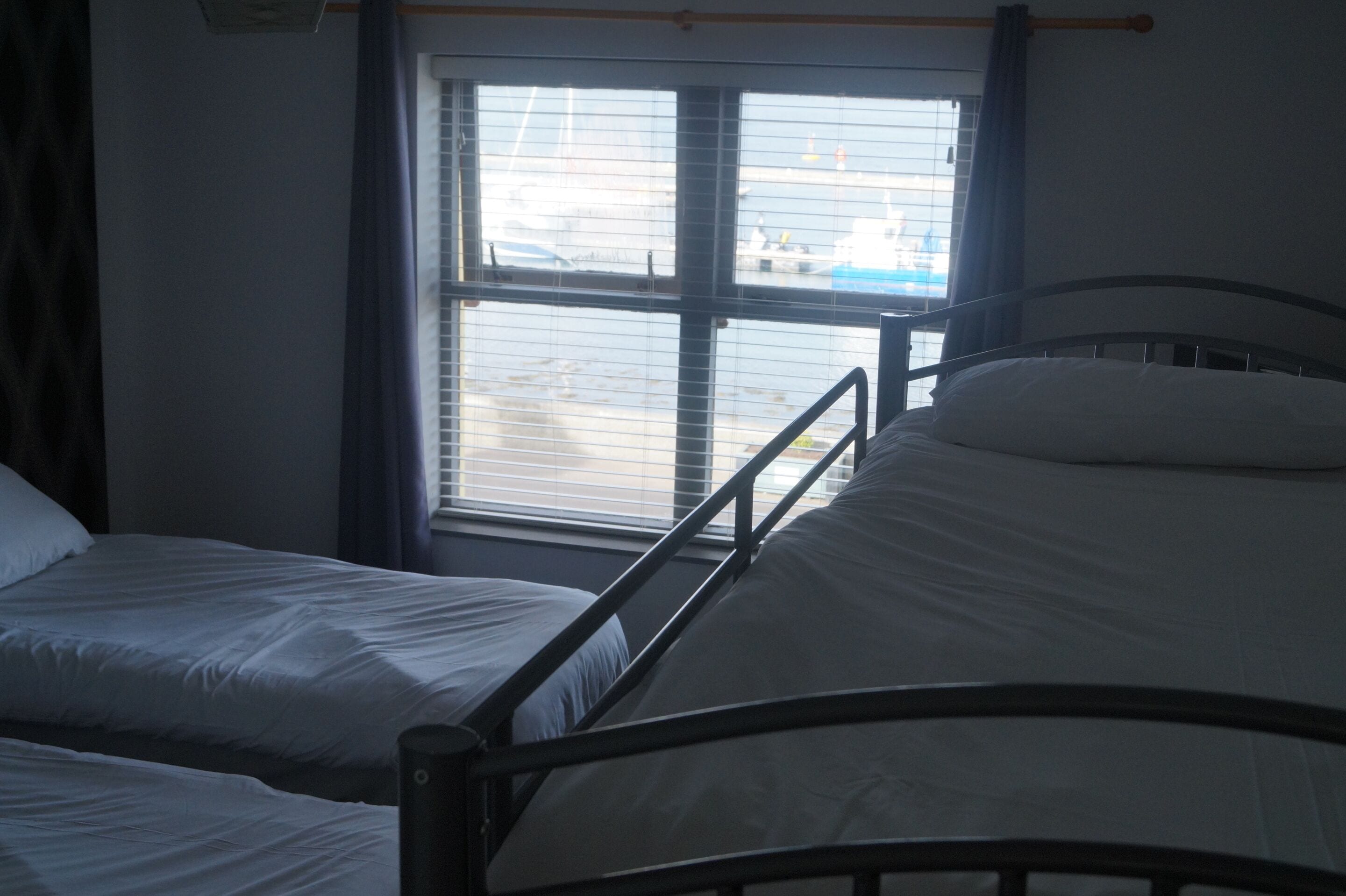A room with two single beds, a window with blinds, and a metal bed frame.