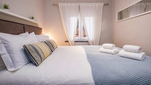 A neatly made bed with white linens and a patterned pillow, a bedside table with a lamp, and a window with sheer curtains.