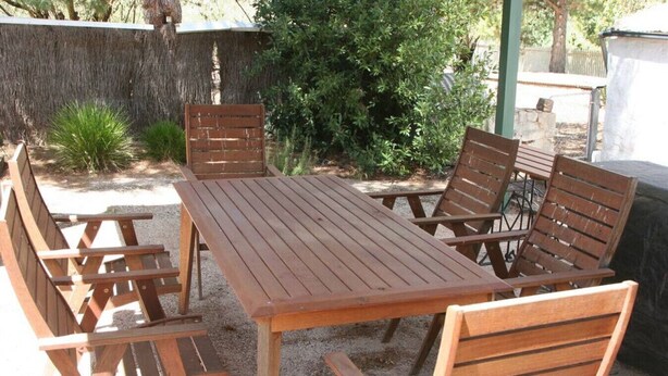 A wooden table and chairs set outdoors with a gravel ground, surrounded by greenery and a stone wall. - Miss Mabel Cottage · Burra - Historic (Burra)