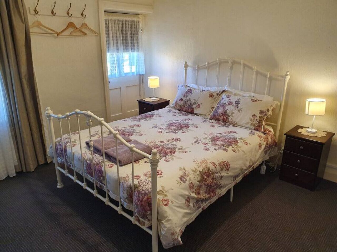 A single bed with a metal frame, a floral bedspread, two bedside tables with lamps, a window with curtains, and a coat rack with hangers.