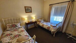 A bedroom with two single beds, a bedside table with lamps, a small desk, and a window with curtains. - Miss Mabel Cottage · Burra - Historic (Burra)