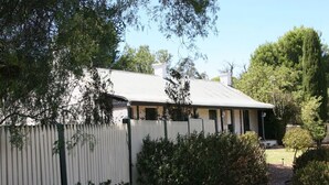 A single-story house with a gabled roof, surrounded by a white picket fence and dense foliage. - Miss Mabel Cottage · Burra - Historic (Burra)