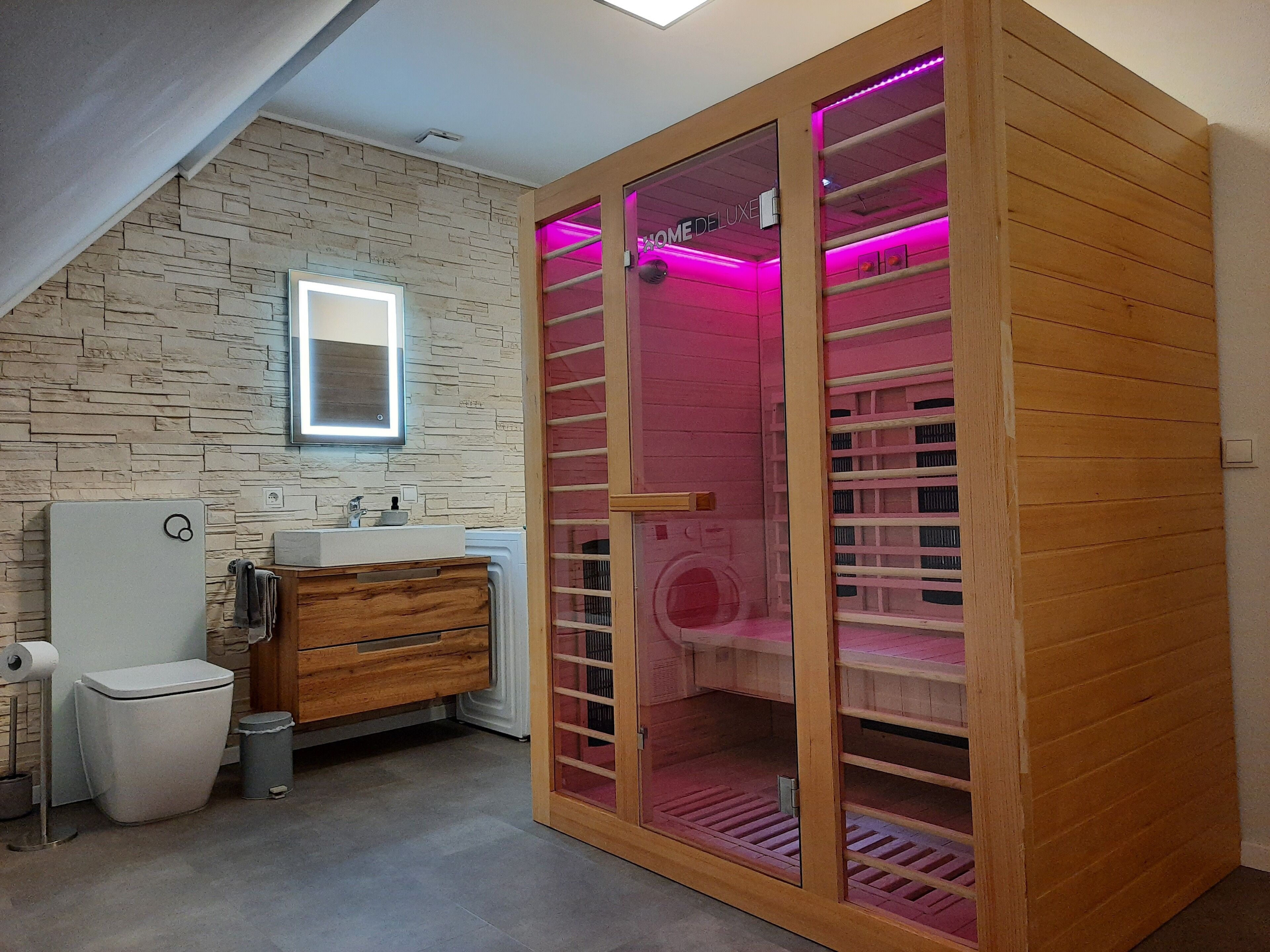 Een badkamer met een stenen muur, een houten kast, een toilet en een douchecabine met glazen wanden en roze verlichting.
