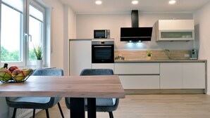 A modern kitchen with a built-in oven, microwave, and a flat-screen TV. There is a dining table with chairs and a fruit bowl.