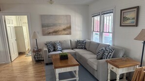 A living room with a sofa, coffee table, and side tables, a painting on the wall, and a door leading to another room.