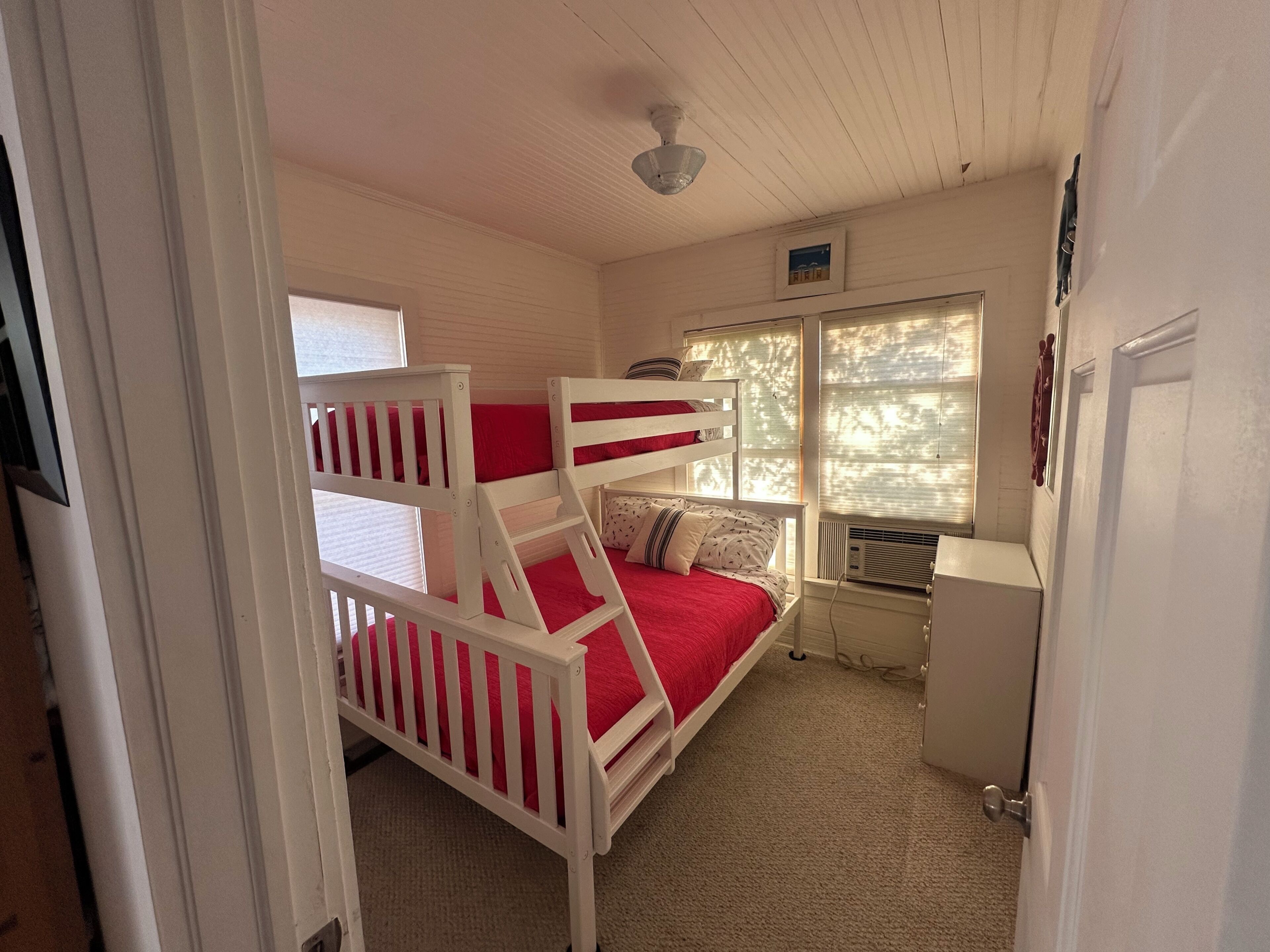 A bunk bed with red bedding, a white ceiling fan, a window with blinds, and a small cabinet.