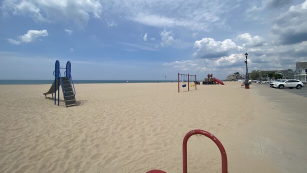 A sandy beach with playground equipment including a slide and swings, a red lamp post, and a parking area with cars.