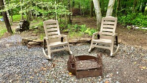 Two rocking chairs and a fire pit in a forested area with gravel ground.