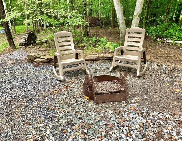 Two rocking chairs and a fire pit in a forested area with gravel ground.