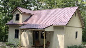 A small, single-story cabin with a sloped metal roof, a porch with two wooden chairs, and a gravel driveway.