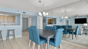 A modern kitchen with a central island, a dining area with blue upholstered chairs, and a large mirror reflecting the space. - Long Beach 2-301 - 2322067 (Panama City Beach)