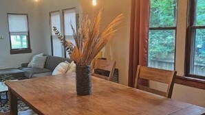 Un coin repas avec une table en bois, des bancs et un vase contenant des plantes séchées. Plusieurs fenêtres sont présentes, ornées de rideaux, et un canapé est visible au fond.