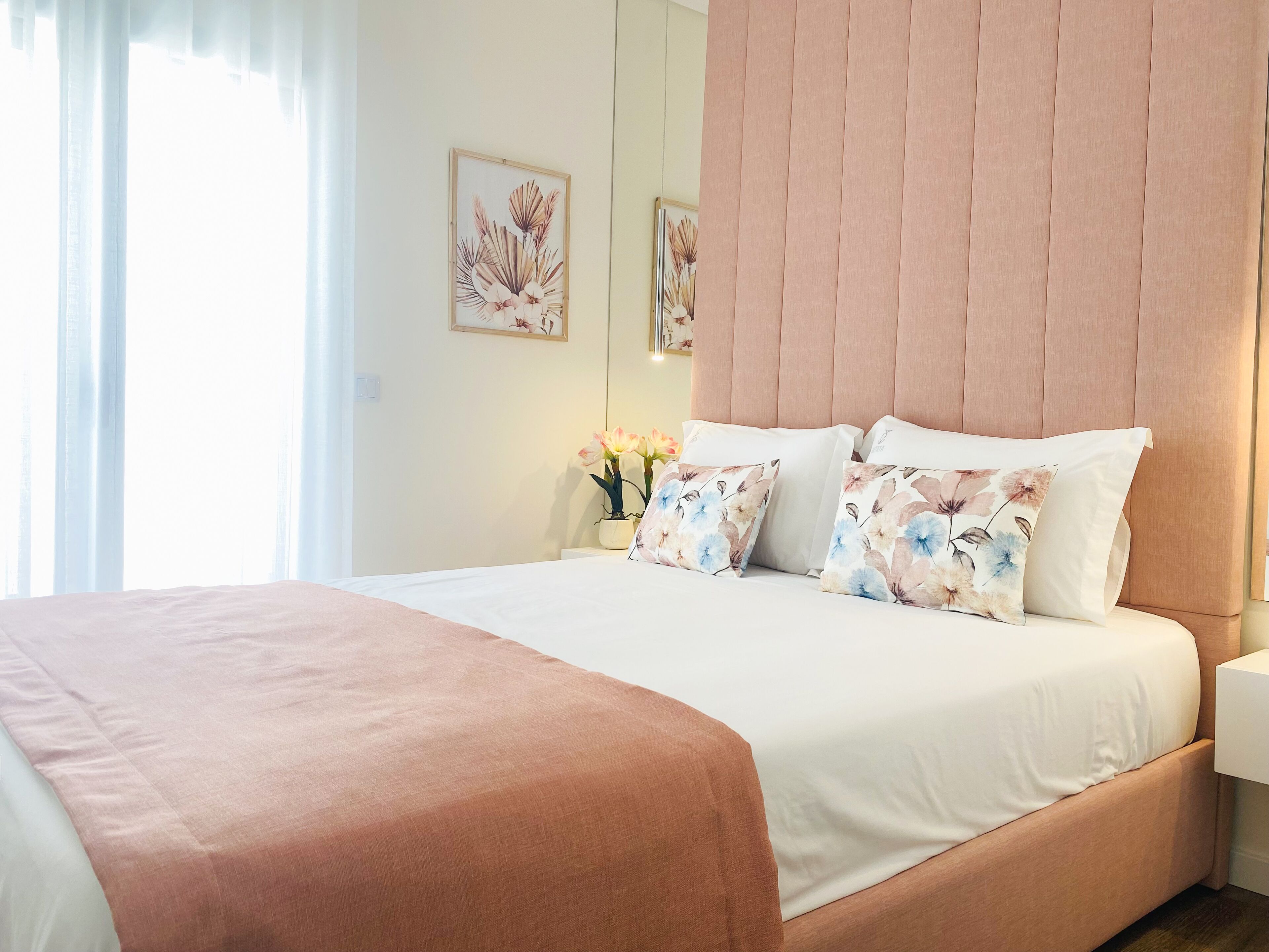 Ein ordentlich bezogenes Bett mit einem rosa Kopfteil, weißer Bettwäsche und Kissen mit Blumenmuster.
