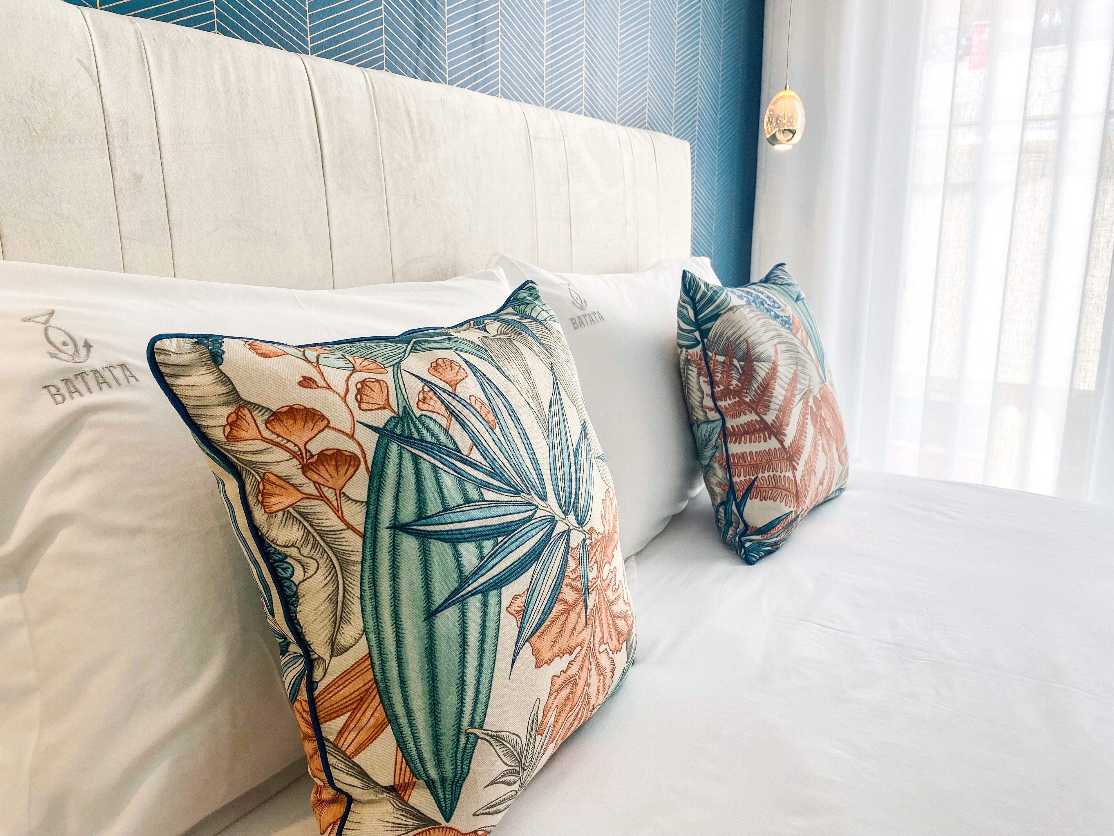 Un lit avec deux coussins décoratifs ornés de motifs de feuilles tropicales et sur lequel l’un des coussins porte le mot « BATATA ».