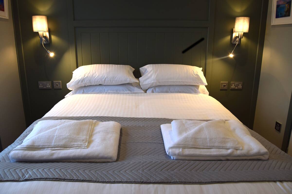 A neatly made bed with white linens, two towels folded on the bed, and wall-mounted lamps on either side.
