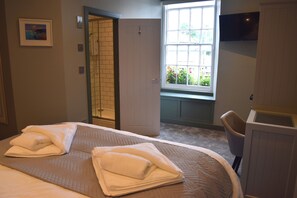 A hotel room with a bed, a window with a view of greenery, a television mounted on the wall, and a bathroom with a shower.