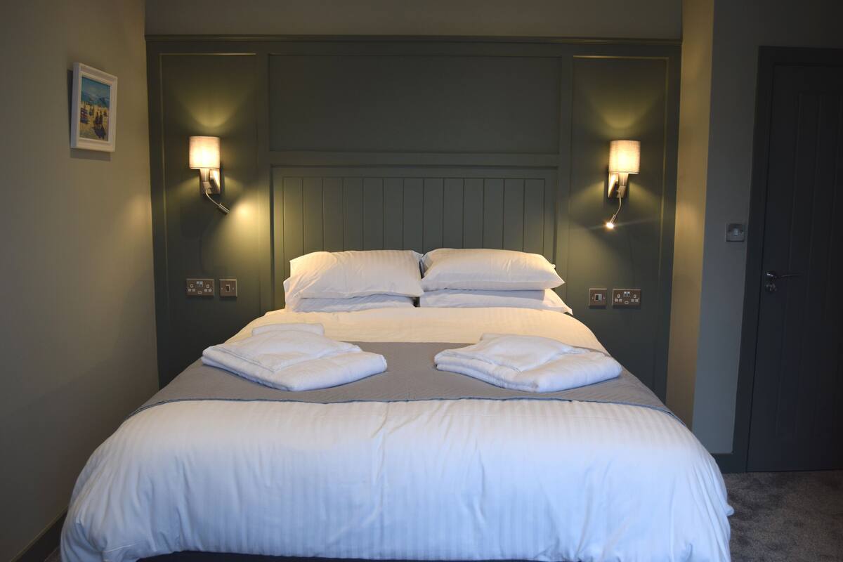 A neatly made bed with white linens and pillows, two wall-mounted light fixtures, and a door to the right.