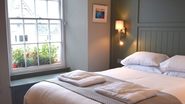 A neatly made bed with white linens and pillows, a window with a view of buildings and flowers, and a wall-mounted light fixture. - Crown Inn (Penzance)