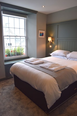 A neatly made bed with white linens and pillows, a window with a view of buildings and flowers, and a wall-mounted light fixture. - Crown Inn (Penzance)