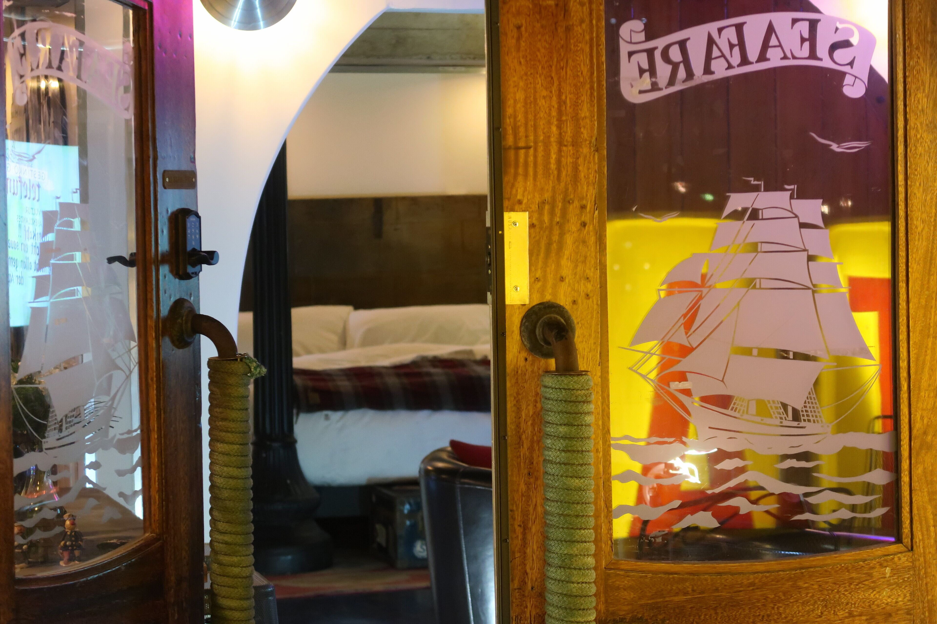 Una habitación con una cama, una puerta de madera con un adhesivo de barco y un espejo que refleja la habitación.