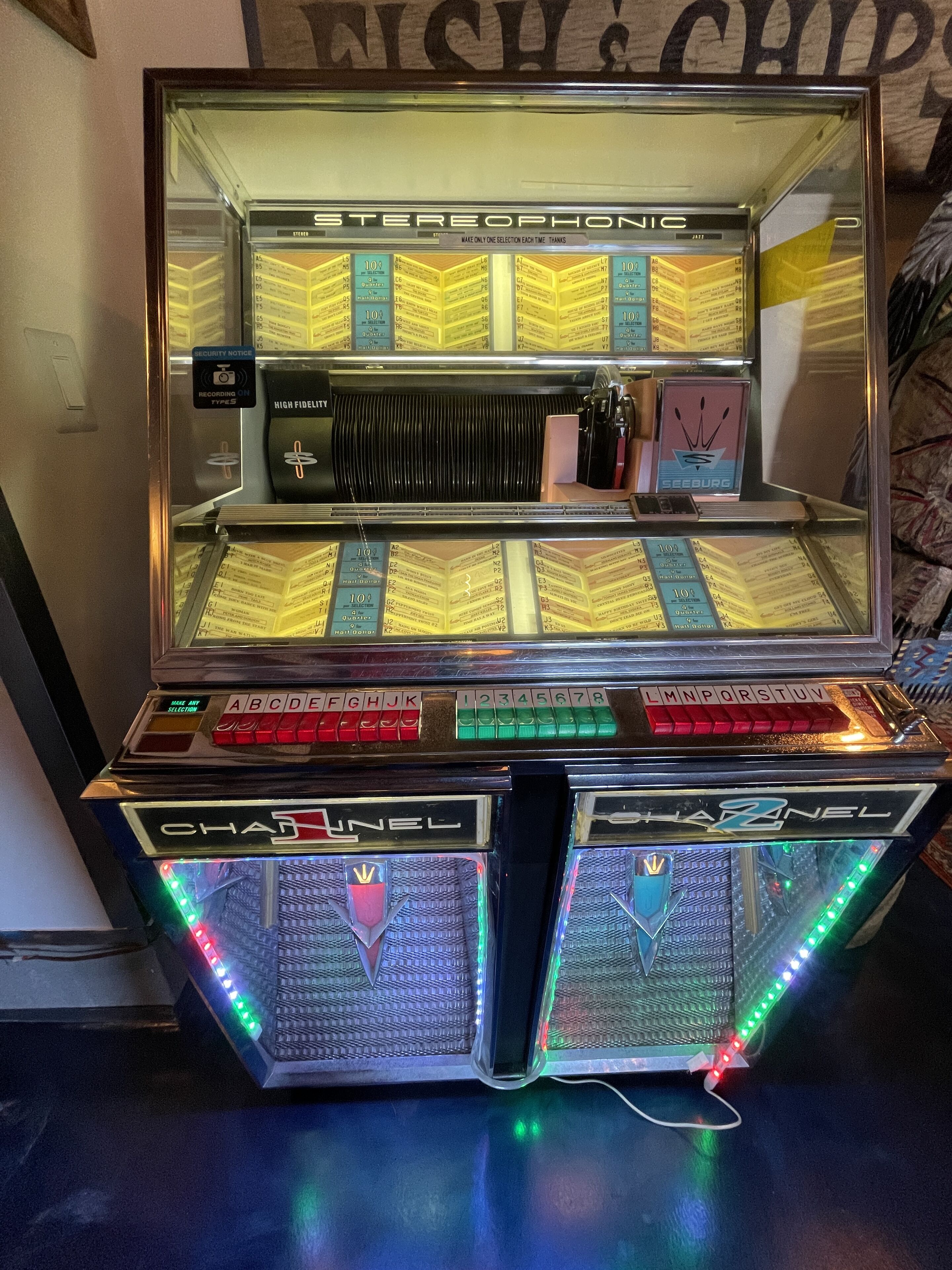 Ein Musikautomat mit einer Auswahl an Schallplatten, einem Sicherheitshinweis und dem Markennamen 'SEEBURG'.
