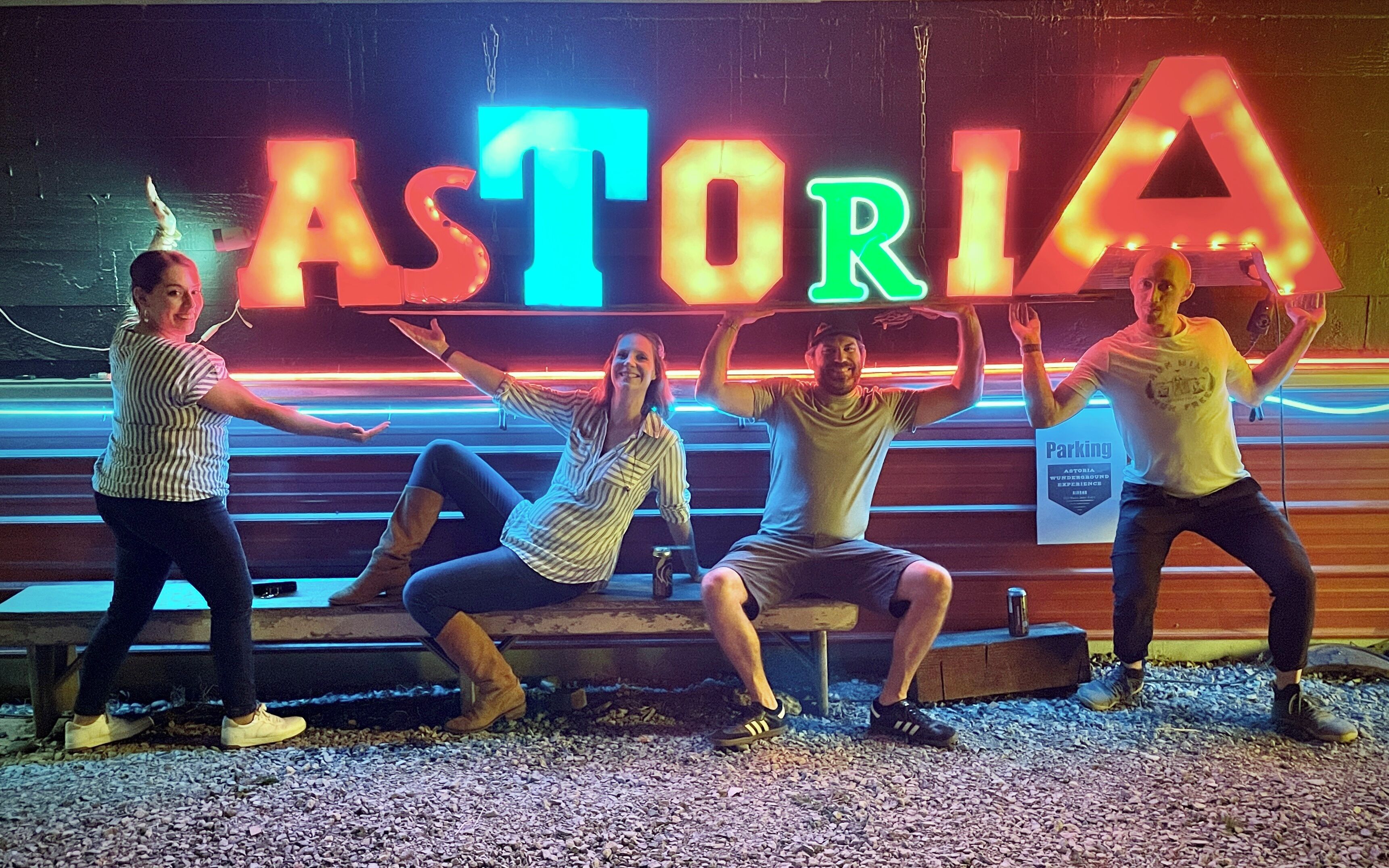 Un grupo de personas posando frente a un letrero de neón con la palabra 'ASTORIA' en una tipografía estilizada.