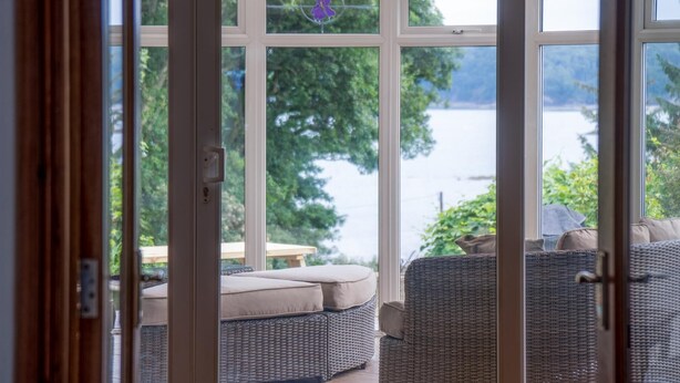 A view through a glass door to a patio area with wicker furniture and a view of a lake. - Sea View - 2 Bedroom Cottage - Wisemans Bridge - Saundersfoot (Saundersfoot)