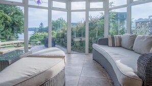 A conservatory with a glass roof, wicker furniture, and a view of a lake. - Sea View - 2 Bedroom Cottage - Wisemans Bridge - Saundersfoot (Saundersfoot)