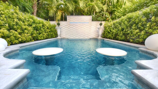 Une piscine rectangulaire avec deux plateformes circulaires blanches au centre, entourée d’un mur de pierre et d’une végétation luxuriante.