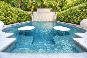 A rectangular swimming pool with two white circular platforms in the center, surrounded by a stone wall and lush greenery. - The Queens Villa (Key West)