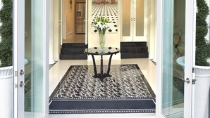 Une entrée imposante, avec un tapis aux motifs noir et blanc, un lustre et une vue sur l’intérieur à travers des portes vitrées.