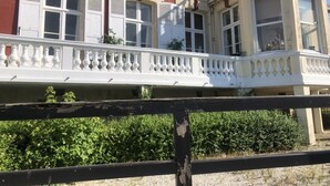 Un edificio con un balcón y barandilla blanca, varias ventanas con contraventanas y una sección en esquina con balcón.