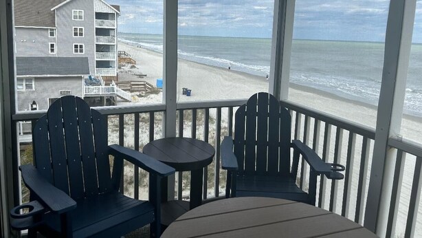 Un espace extérieur abrité avec deux fauteuils Adirondack et une petite table, donnant sur une plage avec un bâtiment en arrière-plan.