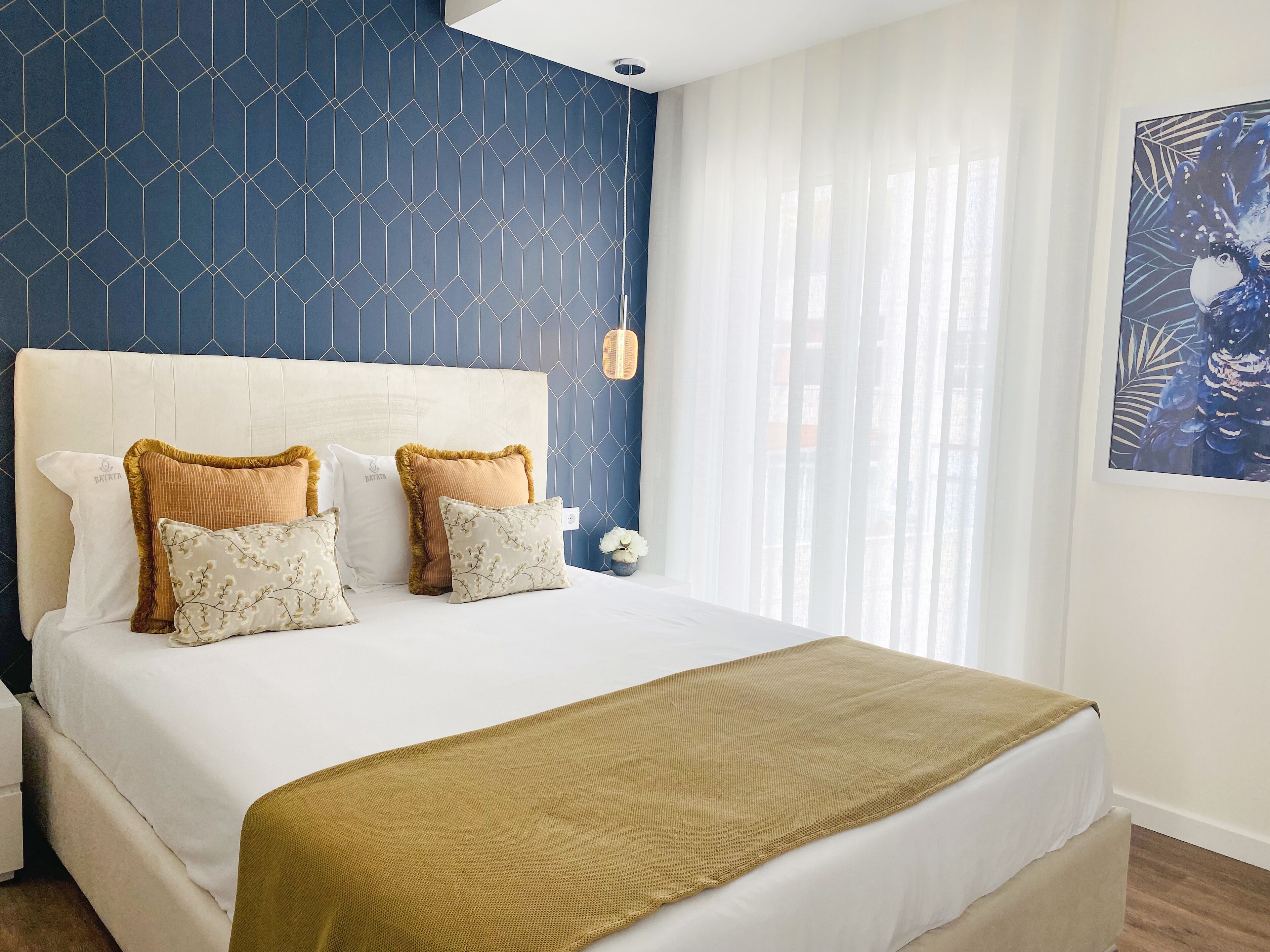 Een netjes opgemaakt bed met een wit hoofdbord en twee kussens, een beige plaid en een muur met een geometrisch patroon.