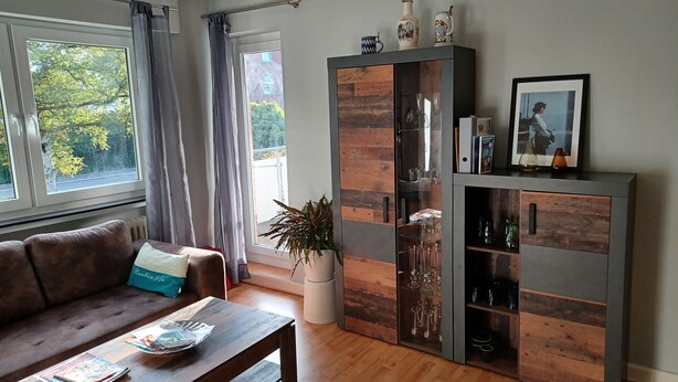 Una sala de estar con un sofá de cuero marrón, una mesa de centro de madera con revistas, un aparador de vidrio con objetos decorativos y vistas a árboles a través de una ventana.