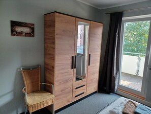 Una habitación con armario de madera, una silla de mimbre, una cama con colchón y almohadas blancas, y una ventana con vistas al verdor exterior.