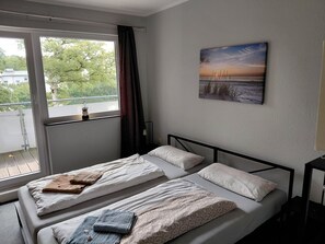 A hotel room with two beds, a window with curtains, a painting on the wall, and a view of trees and buildings outside.