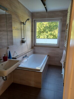 A modern bathroom with a bathtub, a sink, a shower, and a window providing a view of greenery outside.