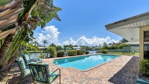 Una piscina con patio de ladrillo, sillas de descanso y vistas a una zona residencial con casas y una masa de agua.