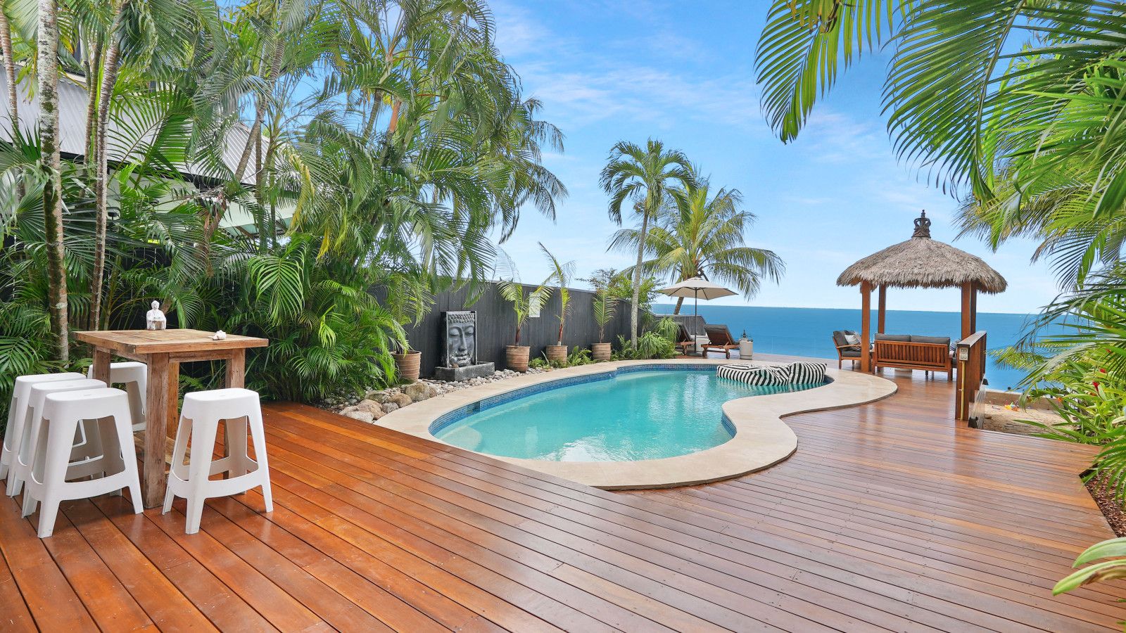 Um deck de madeira com piscina, cadeiras de descanso e um gazebo de palha com vista para o oceano.