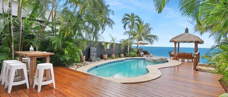 Um deck de madeira com piscina, cadeiras de descanso e um gazebo de palha com vista para o oceano.