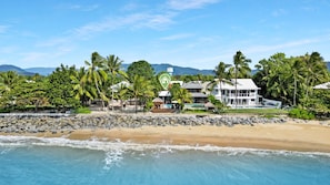 A beachfront property with a rocky shoreline, palm trees, and a large house with multiple levels and a pool.