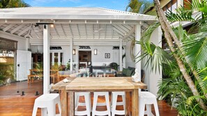 Un espacio de sala comedor de concepto abierto con una mesa de madera y sillas blancas, rodeado de plantas y una estructura blanca en el techo.