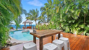 Eine Holzterrasse mit Pool, umgeben von tropischen Pflanzen und Palmen, mit Blick auf den Ozean.