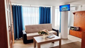A living room with a sofa, a coffee table with a vase of flowers, a flat-screen TV mounted on the wall, and a kitchenette with a microwave. - Hotel Capitol Eforie Sud (Eforie)