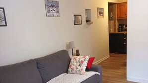 Un salón con un sofá gris, una manta blanca y cojines estampados. Se observan obras de arte en la pared y una lámpara. Se ve una cocina al fondo.